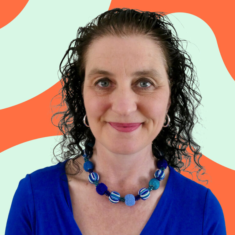 A smiling middle-aged woman with curly hair, wearing a blue top and a colorful beaded necklace, stands against a white background with an abstract orange design.