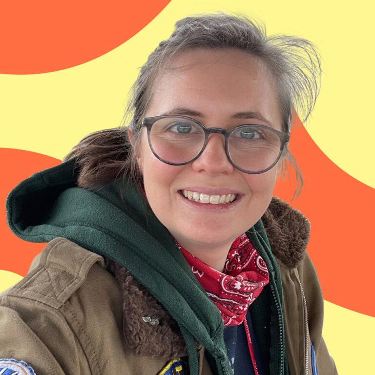 A woman with glasses, smiling at the camera, wearing a green bomber jacket and a red bandana. the background is a bright abstract pattern with yellow and orange swirls.