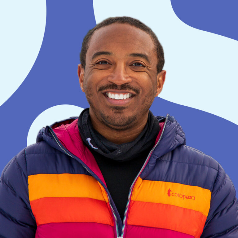 A man with a warm smile, wearing a colorful striped puffer jacket, standing in front of a blue background with a white abstract design.