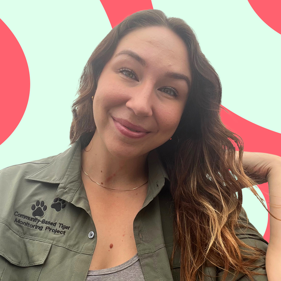 A smiling woman with long brown hair wearing a green shirt that has a logo saying "community-based tiger monitoring project" on it, with a pink circular graphic in the background.