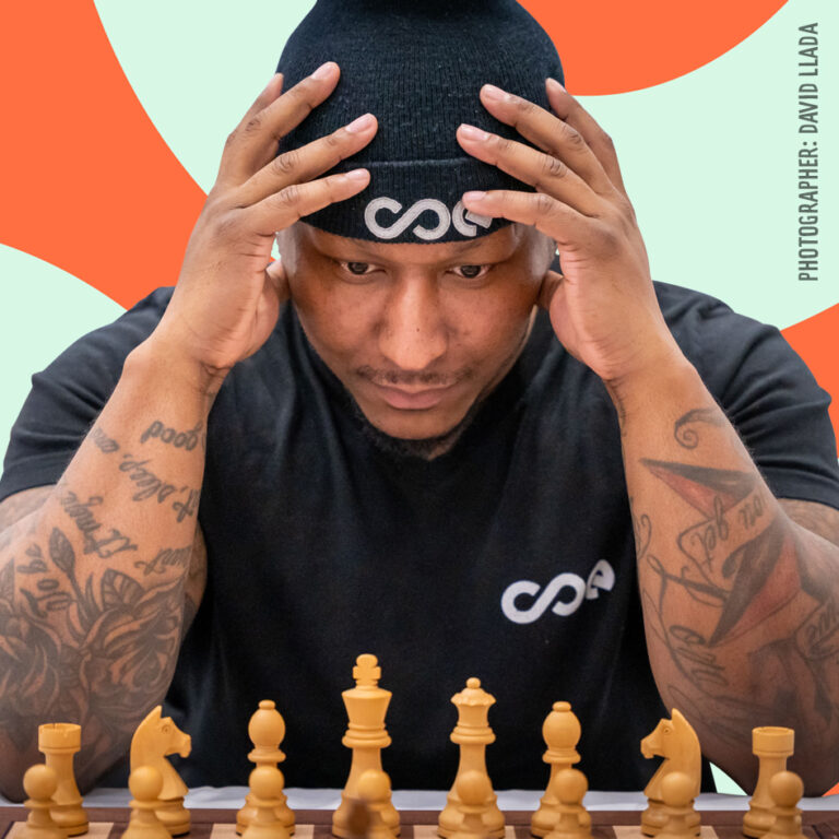 Man with tattoos and beanie focuses on a chessboard, hands on head, against an orange and mint green background.